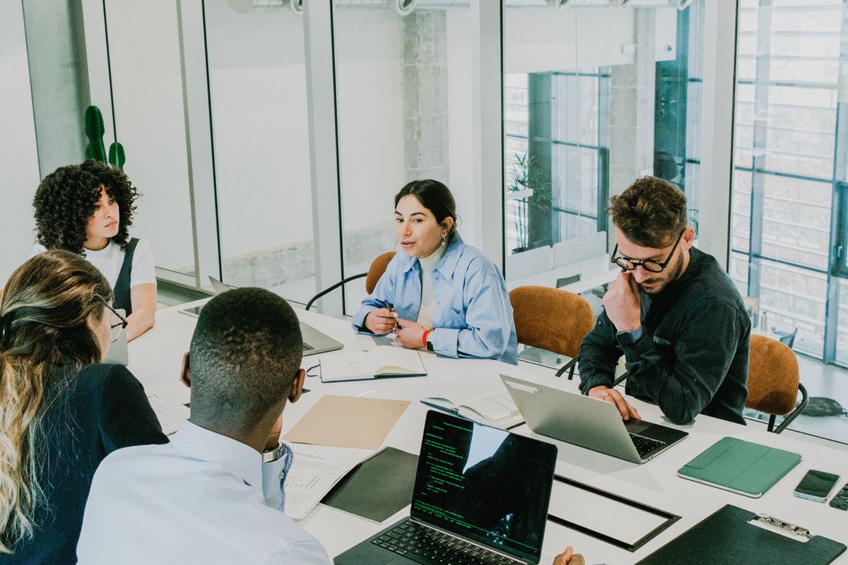 Team meeting in a bright glass office, people discussing around laptops and notebooks, with a focused, collaborative mood.